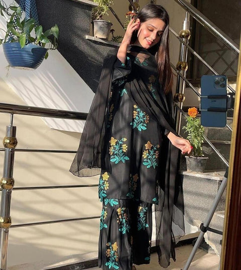 Woman wearing black floral suit set with dupatta, posing indoors in natural light
